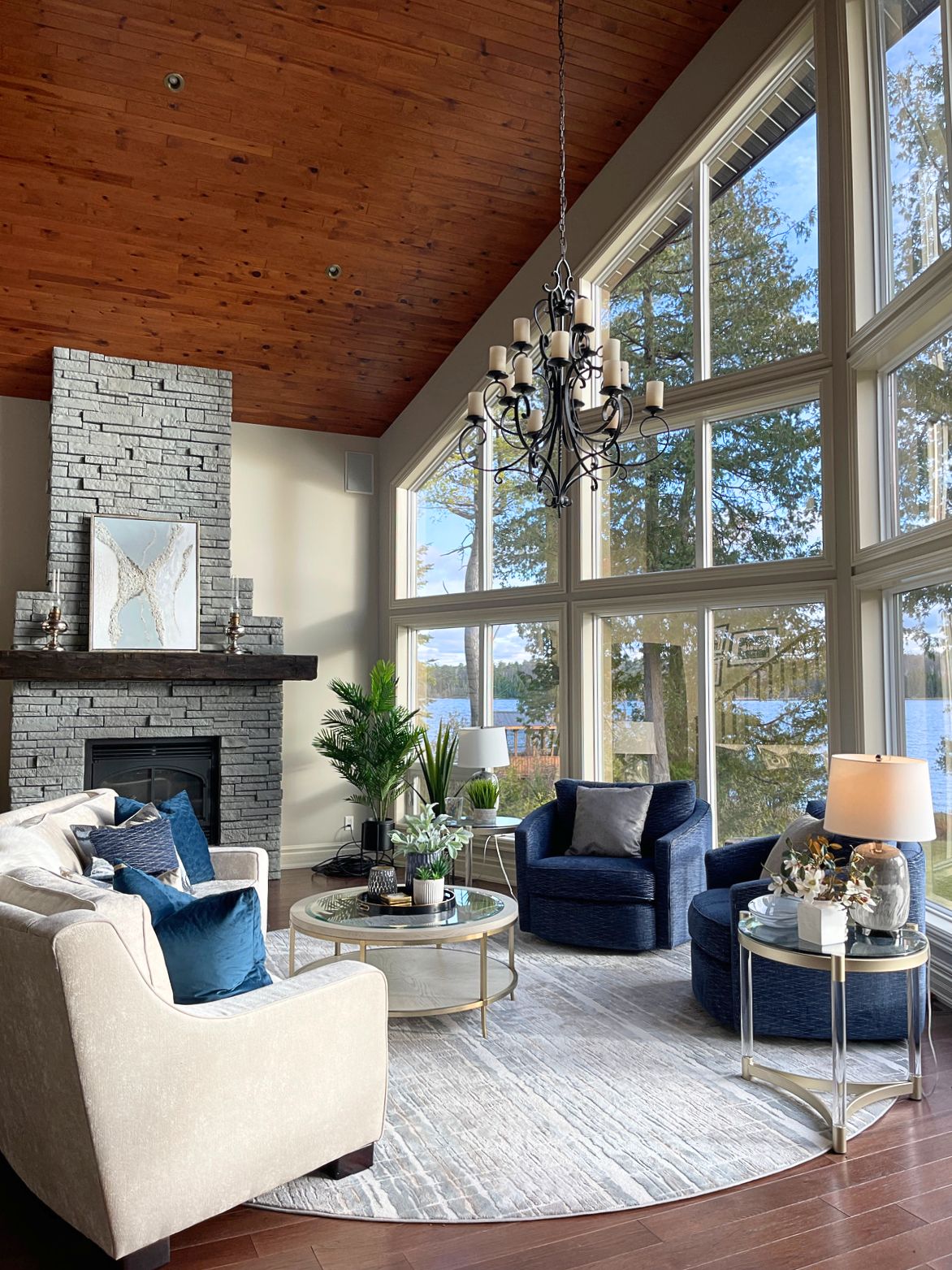 Bright contemporary great room in White Lake featuring floor-to-ceiling lakeview windows, a dark stone fireplace, cream curved sofa, navy accent chairs, and layered modern styling.