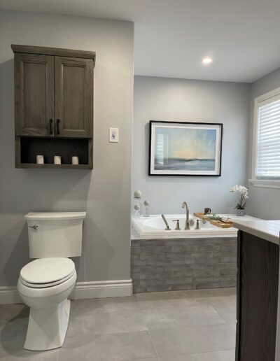 Modern farmhouse bathroom renovation with tiled tub surround, neutral grey walls, framed artwork above tub, and wood wall cabinet over toilet