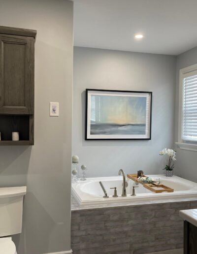 Modern farmhouse bathroom renovation with tiled tub surround, neutral grey walls, framed artwork above tub, and wood wall cabinet over toilet