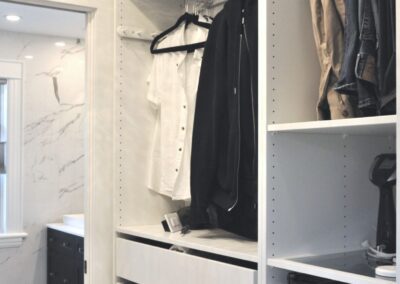 Custom walk-in closet with white built-in cabinetry, drawers, hanging storage, and pendant lighting, with a view into a marble-tiled ensuite bathroom