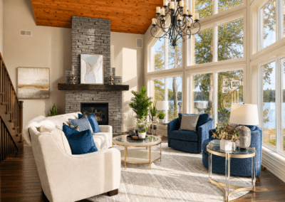 Elegant White Lake great room with soaring lakeview windows, charcoal stone fireplace, cream curved seating, navy swivel chairs, and layered contemporary décor.