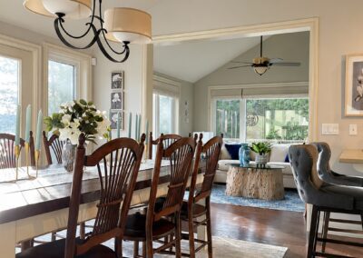 Bright and airy open-concept dining and sitting area in a White Lake lakeside home featuring vaulted ceilings, dark wood flooring, a farmhouse dining table, soft neutral textiles, contemporary lighting, and views into a cozy sitting room with blue accents and natural textures.
