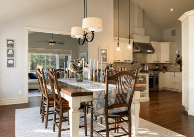 Contemporary lakeside dining room in White Lake featuring a long farmhouse dining table, layered neutral textiles, soft lighting, warm wood flooring, and open views into the adjoining kitchen and sun room.