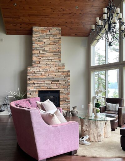 Original White Lake great room with pink seating, light stone fireplace, vaulted wood ceiling, and large windows before redesign.