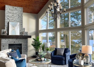 Bright contemporary great room in White Lake featuring floor-to-ceiling lakeview windows, a dark stone fireplace, cream curved sofa, navy accent chairs, and layered modern styling.