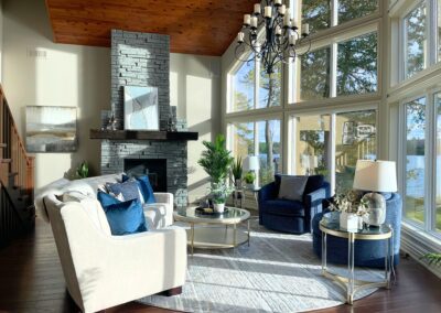 Modern lakeside great room in White Lake featuring a dark stone fireplace, cream curved seating, blue chairs, and dramatic floor-to-ceiling windows.