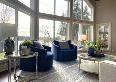 Bright contemporary White Lake great room with navy chairs, layered neutral rug, oversized windows, and lakefront views.